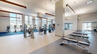 Located at 880 Pacific Street in Stamford, Connecticut, this generously proportioned fitness center spans nearly 3,000 square feet on a single floor, offering a comprehensive and well-appointed workout facility. The space is defined by soaring ceilings, large floor-to-ceiling windows that flood the interior with natural light, and sweeping views of the surrounding Stamford skyline. Linear pendant lighting with warm wood accents complements the modern, neutral color palette throughout.

The main fitness floor is thoughtfully divided into distinct workout zones. A dedicated cardio area, finished with vibrant blue carpet, houses a lineup of treadmills, ellipticals, stationary bikes, and other aerobic machines positioned to take full advantage of the expansive window views. Adjacent to this, a strength training zone features a wide selection of cable machines and selectorized equipment arranged across light-toned flooring for ample circulation space.

A separate free weights area is outfitted with power racks, barbells, weight plates, and a full dumbbell rack, all set against floor-to-ceiling mirrors that enhance the sense of space. A glass-walled studio provides a dedicated space for functional training, complete with TRX suspension systems, stability balls, and open floor space. A sleek glass-partitioned room visible from the main floor adds further versatility to the facility, rounding out this thoroughly equipped fitness destination.