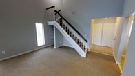 This two-story residence makes an immediate impression with its dramatic vaulted ceilings in the main living area, creating a strong sense of vertical space and openness. The focal point of the room is a well-appointed staircase featuring a dark wood handrail contrasted against crisp white balusters and risers, lending a clean, classic aesthetic. Built-in shelving beneath the staircase provides practical storage while maintaining a tidy appearance.

The living area is finished with neutral beige carpeting, which adds warmth underfoot, while the walls are painted in a soft blue-grey tone that complements the light, airy atmosphere. A window fitted with white blinds allows natural light to filter into the space without sacrificing privacy.

Adjacent to the main living area, a doorway reveals an additional room featuring light hardwood or laminate flooring and a set of bifold closet doors, suggesting use as a bedroom or flex space. The consistent use of white trim, neutral wall tones, and clean finishes throughout gives the space a cohesive, move-in-ready feel suited to a variety of living arrangements.