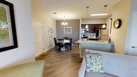 This single-story residence offers a thoughtfully designed open-concept layout that seamlessly connects the living, dining, and kitchen areas. The living room features wood-look flooring, a fireplace with a wall-mounted TV above, a ceiling light fixture, and a front entry door with transom windows that invite natural light and outdoor views. The adjacent dining area is defined by a stylish chandelier, a gray accent wall, and space for a round dining table, creating an intimate yet functional setting.

The kitchen is well-equipped with rich dark cabinetry, granite countertops, stainless steel appliances including a refrigerator and range, and a peninsula with pendant lighting that separates the kitchen from the main living space. A double-door pantry provides generous storage.

The residence includes multiple bedrooms, each finished with plush carpeting, ceiling fixtures, and windows offering outdoor views. The primary bedroom features dual nightstands and a closet, while additional bedrooms include closet storage and space for a dresser and seating.

Two bathrooms serve the home. One features a glass-enclosed tiled shower, granite vanity, and dark-framed mirror. The second includes a shower-tub combination and matching granite countertop, both finished with wood-look flooring and neutral tones throughout.