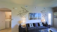 This single-floor space showcases a thoughtfully designed living area that blends contemporary style with natural elements. The living room serves as the focal point, featuring a dark leather sofa adorned with a mix of patterned and solid decorative pillows in muted tones of grey, purple, and beige. A glass-topped coffee table adds a sleek, modern touch to the seating arrangement.

The walls are finished in a soft, neutral tone that provides a clean backdrop for the room's artistic décor. Dried branch arrangements are mounted directly onto the walls, creating an organic, sculptural display that draws the eye and adds texture to the space. A large canvas print depicting a serene forest scene in cool blue and earthy tones serves as a striking focal point above the sofa.

Ambient lighting is provided by warm-toned table lamps with woven shades, positioned on a dark wood nightstand. The ceiling features standard HVAC venting, and a smoke detector is visible, indicating functional, well-maintained infrastructure. Overall, the space conveys a calm, nature-inspired aesthetic with modern furnishings and deliberate decorative choices.