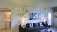 This single-floor space showcases a thoughtfully designed living area that blends contemporary style with natural elements. The living room serves as the focal point, featuring a dark leather sofa adorned with a mix of patterned and solid decorative pillows in muted tones of grey, purple, and beige. A glass-topped coffee table adds a sleek, modern touch to the seating arrangement.

The walls are finished in a soft, neutral tone that provides a clean backdrop for the room's artistic décor. Dried branch arrangements are mounted directly onto the walls, creating an organic, sculptural display that draws the eye and adds texture to the space. A large canvas print depicting a serene forest scene in cool blue and earthy tones serves as a striking focal point above the sofa.

Ambient lighting is provided by warm-toned table lamps with woven shades, positioned on a dark wood nightstand. The ceiling features standard HVAC venting, and a smoke detector is visible, indicating functional, well-maintained infrastructure. Overall, the space conveys a calm, nature-inspired aesthetic with modern furnishings and deliberate decorative choices.