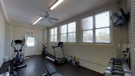 This fitness room is housed within a single-story structure and is defined by its painted white brick walls, which lend the space a clean, utilitarian character. A row of large windows lines one wall, allowing natural light to fill the room and offering views of the surrounding outdoor greenery, creating a bright and open atmosphere despite the room's modest scale. The dark rubber flooring provides a durable, slip-resistant surface well-suited for exercise. Overhead, a ceiling fan aids in air circulation during workouts, complemented by fluorescent strip lighting that ensures consistent illumination. The space is equipped with a treadmill, a stationary bike, and a weight bench, covering both cardio and strength training needs. A clearly marked exit door is positioned along one wall, contributing to the room's safety and accessibility. Overall, the space presents a no-frills, practical fitness environment with adequate ventilation, natural light, and essential equipment for a well-rounded workout routine.