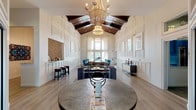 This sophisticated single-story common space presents a striking architectural design centered around dramatically vaulted ceilings with rich dark wood beams that contrast beautifully against the crisp white interior. Elegant chandelier lighting cascades from the ceiling's peak, illuminating the expansive open-plan layout below.

The space is unified by light hardwood flooring and detailed wall paneling featuring classic wainscoting and inset framing throughout. Multiple seating arrangements are thoughtfully distributed across the floor plan, incorporating plush gray sofas, vibrant teal accent chairs, and curated display shelving. A round statement table anchors the entry area, setting a refined tone upon arrival.

A built-in bar counter with bar-height stools provides a casual gathering spot, accented by decorative tile work and pendant lighting. Curated artwork in coordinating blue and neutral tones lines the paneled walls, contributing to a cohesive and polished aesthetic.

At the rear, oversized arched windows and glass doors flood the interior with natural light and frame direct views of an outdoor pool and fountain beyond. The combination of architectural detailing, intentional furnishings, and seamless indoor-outdoor connectivity creates a welcoming, upscale communal atmosphere throughout.