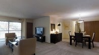 This single-floor residential space offers an open-concept design that seamlessly connects the living and dining areas, creating a cohesive and functional layout. The living area is anchored by a media console and provides ample room for comfortable seating, with neutral-toned walls and plush wall-to-wall carpeting that lend the space a warm, welcoming ambiance. A low partition wall subtly defines the transition between the living and dining zones without interrupting the open feel of the interior. The dining area accommodates a full table and chair set, illuminated by a ceiling-mounted light fixture that adds a soft, inviting glow. A dark wood dresser or buffet with decorative accessories and framed wall art adds character and a sense of style to the shared space. Natural light filters in through windows, complementing the interior lighting. A doorway visible in the background suggests additional rooms beyond, contributing to the overall sense of a well-organized, flowing floor plan. The consistent use of neutral tones, dark wood furnishings, and soft carpeting throughout gives the space a cohesive and tastefully appointed aesthetic.