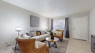 This single-floor residence offers a thoughtfully designed layout with a warm, contemporary aesthetic carried consistently throughout. The living room serves as the heart of the home, featuring light-toned tile flooring, soft neutral walls, and a large window dressed with elegant gray curtains that invite generous natural light. A decorative mandala wall piece and carefully chosen furnishings lend the space a curated, welcoming ambiance.

The dining area is anchored by a stylish chandelier overhead and a large mirror that amplifies the sense of space, creating an intimate yet open setting ideal for entertaining. Adjacent to it, the kitchen is outfitted with crisp white cabinetry, granite-style countertops, and black appliances, offering a functional and visually striking contrast.

The bedroom is a restful retreat, complete with a ceiling fan, natural light filtering through patterned curtains, and a spacious layout accommodating a king-sized bed with a tufted headboard flanked by wall-mounted sconces. A dedicated dressing area with a walk-in closet and console provides additional organization and storage.

The bathroom features a white vanity with granite countertop, a large mirror, and tile flooring, maintaining the clean and cohesive design language found throughout the residence. Light tile flooring runs seamlessly across the home, unifying each space with a bright, airy feel.