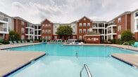 Located at 504 Boxelder Way in Greenville, North Carolina, this outdoor amenity space showcases a generously sized, resort-style swimming pool as its centerpiece. The pool features a distinctive cross-shaped layout with easy-access entry steps and clear blue water, making it both visually striking and functional. The surrounding concrete sundeck is expansive, providing ample room for relaxation and social gathering.

The deck is well-appointed with a variety of seating options, including Adirondack chairs in vibrant teal, cushioned chaise lounges, and wave-shaped loungers, offering residents multiple ways to enjoy the outdoor environment. Mature flowering trees and manicured hedgerows line the perimeter, adding natural color and greenery to the setting.

A dedicated pool house structure, constructed in matching brick, is situated at one end of the deck and includes posted pool rules and safety signage. The entire pool area is enclosed by a black iron fence, ensuring both security and a clean aesthetic boundary.

The space is framed on multiple sides by multi-story residential buildings featuring classic red brick facades, neutral siding accents, and private balconies. The architectural consistency between the pool house and surrounding buildings creates a cohesive, well-planned community atmosphere ideal for outdoor recreation and leisure.