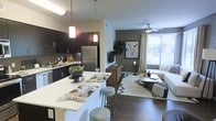 This single-floor residence showcases an open-concept layout that seamlessly connects the kitchen and living area, creating a cohesive and functional living environment. The kitchen is outfitted with dark espresso cabinetry, stainless steel appliances, and a tiled backsplash that adds a contemporary edge. A white countertop island with bar seating serves as a central gathering point, illuminated by pendant lighting that adds warmth and visual interest overhead.

The living area flows naturally from the kitchen, featuring wood-look flooring and an accent wall in a deep olive-green tone that anchors the space with a sense of depth and character. A ceiling fan provides both comfort and a finishing architectural touch. Natural light enters generously through a large window dressed with neutral curtains, brightening the space and offering an airy feel. The open layout encourages versatile furniture arrangement, as demonstrated by the comfortable seating area centered around a chevron-patterned area rug and a wooden media console. Together, these elements create a modern, welcoming atmosphere well-suited for everyday living and entertaining.