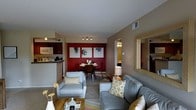 This single-floor residence showcases a thoughtfully designed open-concept layout that seamlessly connects the living, dining, and kitchen areas. The living room is furnished with a comfortable gray sofa and matching armchair, accented by gold and white patterned throw pillows that complement the neutral taupe wall tones. A light wood coffee table anchors the seating area, which sits atop a neutral area rug.

The dining area serves as a focal point, featuring a bold red accent wall adorned with a trio of black-and-white abstract artworks. A round dark wood dining table with matching chairs is centered beneath a track lighting fixture, adding a contemporary touch.

The kitchen is partially visible and includes dark cabinetry, light countertops, a microwave, and a sink with under-cabinet lighting, offering functional workspace. A framed mirror on one wall enhances the sense of depth throughout the space.

Hallways lead to additional rooms, hinting at a well-organized floor plan. Overall, the residence balances modern aesthetics with warm, homey elements, creating a cohesive and comfortable living environment.