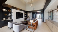 Located in Romeoville, IL, this contemporary single-floor clubhouse is designed with a sophisticated, modern aesthetic that balances bold contrasts with inviting warmth. The main lounge area makes an immediate impression with soaring ceilings, dramatic dark accent beams, and a striking starburst chandelier that anchors the space. A white stacked-stone feature wall frames a large mounted flat-screen TV, flanked by built-in dark shelving with curated décor. Comfortable seating — including a rich cognac leather sofa and soft gray accent chairs — is arranged over patterned area rugs atop light-toned tile flooring, creating a welcoming gathering atmosphere. A corridor with exposed beam detailing and track lighting connects the lounge to additional areas, with glass entry doors drawing in natural light. The full kitchen is outfitted with light wood cabinetry, white subway tile backsplash, stainless steel appliances including a full refrigerator and wall oven, and a generous island with bar seating beneath a row of industrial-style pendant lights. Adjacent to the kitchen, a dedicated dining room features a long dark wood communal table, upholstered chairs, a dramatic dark shiplap ceiling with recessed lighting, and large windows that flood the room with natural light and offer open views of the surrounding landscape.