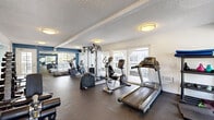This fitness center offers a comprehensive workout environment within a single, open-plan floor. The space is designed to accommodate a wide variety of exercise routines, featuring an impressive array of equipment that includes treadmills, elliptical trainers, a recumbent bike, a multi-station cable machine, a rowing machine, and a dedicated free weights area with dumbbells, kettlebells, and a barbell with weight plates. A stretch and floor exercise zone provides additional versatility for functional training.

The interior is characterized by a textured ceiling with exposed beams and flush-mounted ceiling lights that provide consistent, even illumination throughout. Bold blue accent walls contrast with crisp white surfaces, creating an energetic yet clean aesthetic. Large windows and glass-paned doors line the perimeter, flooding the space with natural light and offering views of an outdoor balcony area beyond. Full-length mirrors are strategically placed to enhance the sense of openness and assist with form during exercise.

Durable rubber flooring covers the entire workout area, providing safety and comfort underfoot. The overall ambiance is practical and motivating, offering users a well-organized, bright, and fully equipped facility suited for a broad range of fitness goals.