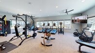 Located at 7135 Mount Zion Circle in Morrow, Georgia, this fitness center offers a well-appointed exercise space designed to support a wide range of workout routines. The single-floor layout is thoughtfully organized to maximize functionality while maintaining an open, comfortable atmosphere.

Large windows line the perimeter of the room, flooding the space with natural light and providing views of the surrounding outdoor areas, including a courtyard and adjacent buildings. Recessed ceiling lights and ceiling fans ensure consistent illumination and airflow throughout.

The cardio zone features multiple treadmills, an elliptical trainer, a stair climber, and a stationary cycling bike equipped with an interactive display screen. The strength training area is equally comprehensive, housing a True multi-station cable machine, a power rack with free weights, a flat bench press, and a full dumbbell rack. A dedicated functional training station includes TRX suspension straps, resistance bands, medicine balls, kettlebells, and a TKO heavy punching bag. A BOSU balance trainer and battle ropes round out the functional fitness offerings.

Wall-mounted mirrors expand the sense of space, while a large flat-screen television adds an entertainment element. The neutral-toned carpeted flooring and light gray walls create a clean, modern aesthetic that makes this fitness center an inviting and practical facility.