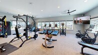 Located at 7135 Mount Zion Circle in Morrow, Georgia, this fitness center offers a well-appointed exercise space designed to support a wide range of workout routines. The single-floor layout is thoughtfully organized to maximize functionality while maintaining an open, comfortable atmosphere.

Large windows line the perimeter of the room, flooding the space with natural light and providing views of the surrounding outdoor areas, including a courtyard and adjacent buildings. Recessed ceiling lights and ceiling fans ensure consistent illumination and airflow throughout.

The cardio zone features multiple treadmills, an elliptical trainer, a stair climber, and a stationary cycling bike equipped with an interactive display screen. The strength training area is equally comprehensive, housing a True multi-station cable machine, a power rack with free weights, a flat bench press, and a full dumbbell rack. A dedicated functional training station includes TRX suspension straps, resistance bands, medicine balls, kettlebells, and a TKO heavy punching bag. A BOSU balance trainer and battle ropes round out the functional fitness offerings.

Wall-mounted mirrors expand the sense of space, while a large flat-screen television adds an entertainment element. The neutral-toned carpeted flooring and light gray walls create a clean, modern aesthetic that makes this fitness center an inviting and practical facility.