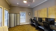 This single-floor space is designed as a functional workspace, highlighted by a row of computer workstations equipped with monitors and ergonomic chairs, making it well-suited for shared or collaborative work. The room features warm cork flooring that adds texture and a natural aesthetic underfoot, complemented by neutral gray walls that create a calm, focused atmosphere.

Crown molding runs along the ceiling perimeter, lending the space a refined architectural detail, while a combination of recessed lighting and a central ceiling fixture ensures even, practical illumination throughout. A window fitted with horizontal blinds allows natural light to filter in, offering a view of surrounding greenery and contributing to an airy feel.

A decorative folding screen partition positioned near the window adds a subtle design element, providing visual separation without enclosing the space. Large corkboard panels mounted on the wall serve both a functional and decorative purpose, offering ample space for notes, documents, or creative planning. A framed piece of wall art adds a personal touch. Overall, the space balances practicality with thoughtful design details.
