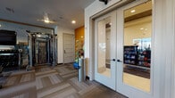 This single-floor fitness center is thoughtfully designed to accommodate a wide range of workout styles and fitness goals. The space is divided into several distinct functional zones, each tailored to a specific type of training. The main gym floor features patterned, multi-tone carpet tiles throughout, providing both durability and a modern aesthetic. Recessed lighting and ceiling fans ensure a comfortable, well-lit environment during workouts.

The cardio zone is outfitted with a selection of Matrix-brand machines, including ellipticals, a stair climber, and additional cardio equipment, all positioned near large windows that allow natural light and outdoor views. Adjacent to this is a strength training area with cable machines, a weight bench, and a rack of dumbbells, complemented by floor-to-ceiling mirrors that enhance the sense of openness.

A dedicated functional fitness area showcases a RAX multi-purpose rig with stall bars, suspension trainers, resistance bands, and a wall-mounted storage rack holding medicine balls and other accessories. This zone also features a ballet barre and mirrored wall, making it suitable for stretching, yoga, or group classes.

A glass-paneled door leads to a separate enclosed room, visible from the main floor, which appears to serve as a private studio or changing area. A vending area with snacks is accessible nearby. Bold accent walls in amber and gray add visual energy throughout, reinforcing the space's active, motivating atmosphere.