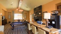 This inviting single-floor amenity lounge is thoughtfully designed to offer both entertainment and relaxation in one cohesive space. The room's most striking feature is a floor-to-ceiling dark slate fireplace wall, accented with warm wood inlays and a bold decorative sculptural element, flanked by wall sconces and framed artwork that add a refined, lodge-inspired aesthetic. Anchoring the center of the room is a full-size pool table set atop a striped area rug, illuminated by a distinctive wrought-iron and alabaster pendant light that serves as a dramatic focal point overhead. Adjacent to the game area, a generous granite-topped bar counter with cream upholstered high-back stools provides a comfortable spot for gathering and socializing. The warm amber and terracotta tones of the walls create an enveloping, welcoming ambiance throughout. A casual seating area with a sofa and soft lighting is tucked near an arched window, allowing natural light to filter in and brighten the space. Recessed lighting and carefully placed lamps complement the overall warm glow of the room. The combination of entertainment features, lounge seating, and distinctive architectural details makes this space well-suited for social gatherings and leisure.