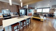 Located at 2710 N Proctor St in Tacoma, Washington, this single-floor community space showcases a thoughtfully designed open-concept layout that seamlessly integrates a full kitchen, multiple dining areas, and comfortable lounge spaces. The interior is finished with warm wood-tone cabinetry, light hardwood-style flooring, and a cohesive contemporary aesthetic throughout.

The kitchen is a central highlight, featuring stainless steel appliances including a French door refrigerator, electric range, and microwave. A large island with a white quartz countertop and undermount sink provides bar-height seating and serves as a natural gathering point. Pendant lighting illuminates the island, while a striking floor-to-ceiling wood-paneled accent wall with an integrated vertical wine rack adds a distinctive design element.

The lounge area offers plush seating arranged around a bold black tile fireplace with an electric insert, creating a warm focal point. A wall-mounted television on a warm wood panel serves an adjacent seating area. The dining zones feature high-top tables with booth-style and chair seating, accented by patterned window treatments and large windows that invite natural light and views of the surrounding Tacoma neighborhood. Lush indoor plants and curated wall art contribute to the space's welcoming, community-oriented ambiance.