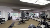 The Stonegate Apartments Fitness Center at 1004 La Bonne Pkwy in Manchester, Missouri is a dedicated, single-floor exercise facility designed to meet a broad range of fitness needs. The space features a generous open floor plan finished with durable, rubber-backed carpet flooring suited for athletic use. Large floor-to-ceiling windows along the exterior walls flood the room with natural light and provide views of the surrounding greenery, creating an inviting workout environment. Full-length wall mirrors further amplify the sense of space and allow users to monitor their form during exercise. Overhead, recessed skylight panels supplement the natural light with bright, even illumination throughout the facility. The cardio section is well-appointed with treadmills, elliptical trainers, and a stationary bike, while a multi-station cable weight machine serves as the centerpiece for strength training. A dedicated free weight area includes a rack of dumbbells and a flat workout bench. Additional equipment such as a rowing machine and exercise ball rounds out the diverse offerings. Practical amenities include a water fountain, a storage cabinet, and posted facility rules. The neutral-toned walls and clean layout contribute to a functional, organized atmosphere suited for both casual and focused training sessions.