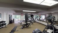 The Stonegate Apartments Fitness Center at 1004 La Bonne Pkwy in Manchester, Missouri is a dedicated, single-floor exercise facility designed to meet a broad range of fitness needs. The space features a generous open floor plan finished with durable, rubber-backed carpet flooring suited for athletic use. Large floor-to-ceiling windows along the exterior walls flood the room with natural light and provide views of the surrounding greenery, creating an inviting workout environment. Full-length wall mirrors further amplify the sense of space and allow users to monitor their form during exercise. Overhead, recessed skylight panels supplement the natural light with bright, even illumination throughout the facility. The cardio section is well-appointed with treadmills, elliptical trainers, and a stationary bike, while a multi-station cable weight machine serves as the centerpiece for strength training. A dedicated free weight area includes a rack of dumbbells and a flat workout bench. Additional equipment such as a rowing machine and exercise ball rounds out the diverse offerings. Practical amenities include a water fountain, a storage cabinet, and posted facility rules. The neutral-toned walls and clean layout contribute to a functional, organized atmosphere suited for both casual and focused training sessions.
