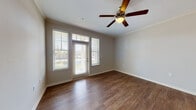 This single-floor residence offers a cohesive and comfortable layout designed for everyday living. The living area serves as the heart of the home, showcasing rich, dark hardwood-style flooring, elegant crown molding, and a ceiling fan with a light fixture that adds both function and style. Large windows with horizontal blinds flank a glass-panel entry door, flooding the space with natural light and providing views of the surrounding exterior. The open-concept design connects the living area to the kitchen, which features dark cabinetry with granite-style countertops, black appliances including a refrigerator and range, and durable wood-look flooring. A louvered door leads to an additional storage or utility area within the kitchen corridor. The bedroom offers plush wall-to-wall carpeting, a ceiling fan, and a bright window that continues the home's emphasis on natural light. The bathroom is appointed with a granite-style vanity countertop, dark wood cabinetry beneath the sink, and a wide mirror that enhances the sense of space. Neutral gray walls throughout the residence provide a calm, versatile backdrop, while consistent trim and molding details lend a polished, unified aesthetic to the entire home.
