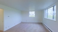 This single-floor space presents a straightforward and functional layout designed for comfortable everyday living. The room visible in the image is generously proportioned, featuring wall-to-wall beige carpeting that adds a sense of warmth underfoot and complements the neutral, light-colored walls throughout. The smooth, flat ceiling enhances the open feel of the room without visual distraction.

Natural light enters through a centrally positioned window on the far wall, offering an outdoor view of a green lawn and surrounding trees, lending a quiet, suburban character to the space. An additional window along the side wall supplements the natural light, keeping the interior bright and airy even in overcast conditions.

Baseboard heating units run along the base of the exterior walls, providing a consistent and efficient heating solution suited to colder climates. Electrical outlets are positioned at standard intervals along the walls, supporting practical furniture arrangements. The overall ambiance is clean, neutral, and move-in ready, making it a versatile space adaptable to a variety of uses and personal styles.