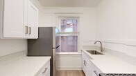 This thoughtfully laid-out single-floor unit makes efficient use of its intimate footprint, offering a cohesive and modern living experience across four distinct areas. The kitchen is outfitted with crisp white shaker cabinetry, brushed nickel hardware, light quartz countertops, a stainless steel single-basin sink, and stainless steel appliances including a refrigerator and gas range. A window fitted with horizontal blinds brings in natural light, enhancing the clean, bright aesthetic. Light hardwood-style flooring runs throughout the main living and sleeping areas, contributing to a warm and unified feel. The bedroom is airy and well-lit, featuring a ceiling fan with integrated light, a mirrored sliding closet door, and a window that allows natural light to filter in. An additional room with dual windows and a wall-mounted heating unit offers flexibility as a secondary living or dining space. The bathroom is finished in an all-white palette, featuring floor-to-ceiling white subway tile in the tub/shower surround, a built-in soaking tub, a wall-mounted vanity with a rectangular sink, brushed gold fixtures, and a sleek illuminated mirror. The overall design is clean, modern, and move-in ready.