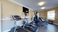 This fitness room offers a well-equipped exercise space designed to support a range of workout routines. The room is finished with warm beige walls, white crown molding, and white baseboards, lending a clean, polished aesthetic that sets it apart from a typical utilitarian gym environment. Flush-mounted ceiling lights provide consistent, even illumination throughout the space.

The cardio section includes a treadmill and an elliptical trainer, both featuring digital display consoles for tracking workout metrics. A flat-screen television is wall-mounted at an elevated position, allowing users to stay entertained during cardio sessions. Adjacent to the cardio equipment is a multi-function cable and weight stack machine, suitable for a variety of strength-training exercises targeting multiple muscle groups.

Along the far wall, a dumbbell rack holds a selection of free weights, complemented by a weight bench, offering additional options for resistance training. The flooring consists of durable, dark rubberized material, providing both cushioning and slip resistance. Overall, the space is thoughtfully arranged to maximize functionality while maintaining a tidy and inviting atmosphere.