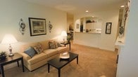 This single-floor residence offers a welcoming and cohesive layout designed for comfortable everyday living. The spacious living room serves as the heart of the home, featuring plush wall-to-wall carpeting in a warm beige tone that adds to the cozy ambiance. Recessed lighting and a table lamp provide a layered lighting scheme, while framed wall art and decorative accents lend a personalized, lived-in character to the space. A comfortable sofa with decorative pillows anchors the seating area, complemented by a dark-finished coffee table that adds a subtle contrast to the light color palette. A graceful arched passageway connects the living area to the kitchen, creating an open and airy feel while maintaining a sense of architectural definition. The kitchen is bright and functional, featuring white cabinetry that extends to the upper walls, a breakfast bar ideal for casual dining, and ample counter space. Adjacent to the living room, a dining area with dark wood chairs offers a dedicated space for meals. Throughout the home, smooth walls painted in soft, neutral tones unify each area, creating a calm and inviting atmosphere that is both practical and aesthetically refined.