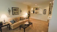 This single-floor residence offers a welcoming and cohesive layout designed for comfortable everyday living. The spacious living room serves as the heart of the home, featuring plush wall-to-wall carpeting in a warm beige tone that adds to the cozy ambiance. Recessed lighting and a table lamp provide a layered lighting scheme, while framed wall art and decorative accents lend a personalized, lived-in character to the space. A comfortable sofa with decorative pillows anchors the seating area, complemented by a dark-finished coffee table that adds a subtle contrast to the light color palette. A graceful arched passageway connects the living area to the kitchen, creating an open and airy feel while maintaining a sense of architectural definition. The kitchen is bright and functional, featuring white cabinetry that extends to the upper walls, a breakfast bar ideal for casual dining, and ample counter space. Adjacent to the living room, a dining area with dark wood chairs offers a dedicated space for meals. Throughout the home, smooth walls painted in soft, neutral tones unify each area, creating a calm and inviting atmosphere that is both practical and aesthetically refined.