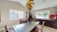 This single-level combined dining room and kitchen space presents a thoughtfully designed interior with a contemporary aesthetic. The vaulted ceiling immediately draws the eye upward, amplifying the sense of openness and serving as an architectural focal point. Suspended from the ceiling's peak is a bold, sculptural gold spiral chandelier that commands attention and anchors the dining area with dramatic visual flair.

The dining area centers around a sleek glass-top table paired with dark-framed chairs upholstered in warm brown tones. Multiple windows along the walls allow generous natural light to fill the space throughout the day, offering views of the surrounding outdoor environment. Neutral gray walls and light wood-tone flooring create a clean, cohesive backdrop throughout.

Dark espresso-finished cabinetry lines multiple walls, providing substantial built-in storage with brushed chrome hardware. A granite-topped built-in workstation with upper shelving and lower drawers adds a functional dimension to the space. French glass doors connect the dining area to adjacent rooms, maintaining an open and airy flow. The overall ambiance is polished and modern, balancing practical storage solutions with refined design details.