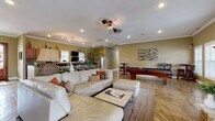 This well-appointed single-floor space offers an open-concept design that seamlessly connects its key areas, creating a welcoming and functional environment throughout. The kitchen is a standout feature, outfitted with rich warm-toned cabinetry, granite countertops, stainless steel appliances, and an extended breakfast bar with bar seating — ideal for casual dining or entertaining. A wall-mounted television and abundant natural light from plantation-shuttered windows add to the kitchen's practicality and comfort.

The expansive main living area is anchored by a striking floor-to-ceiling stone fireplace with a wooden mantle and a wall-mounted flat-screen television, providing a focal point for relaxation. A generously sized sectional sofa and ottoman arrangement creates an inviting lounge setting, while the decorative stamped tile flooring adds visual texture and character throughout the space.

A dedicated game area features a full-size billiards table and pub-height seating, making it well-suited for social gatherings. Crown molding, recessed lighting, ceiling fans with track lighting, and warm neutral wall tones contribute to a cohesive and polished aesthetic. A separate room with natural window views provides a quieter setting for meetings or focused activities, rounding out this versatile and thoughtfully designed space.