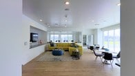 This single-floor space showcases a contemporary open-plan layout that seamlessly integrates a lounge and dining area within a bright, airy environment. The interior is characterized by clean white walls and smooth ceilings fitted with recessed lighting, complemented by warm hardwood-style flooring that extends throughout the space.

The lounge area serves as a focal point, anchored by a bold chartreuse sectional sofa accented with blue and teal throw pillows, arranged atop a chevron-patterned area rug. A round tufted ottoman in blue adds a stylish accent, while a low-profile side table in black provides functional detail. A wall-mounted flat-screen television is set against a textured tile feature wall, adding visual depth to the seating area.

Large floor-to-ceiling windows along the far wall and side elevation flood the interior with natural light and offer open views of the surrounding greenery, enhancing the sense of spaciousness. The dining area to the right features modern tables and chairs in a minimalist style, suitable for casual gatherings. Overall, the space conveys a clean, vibrant, and welcoming atmosphere suited for social or communal use.