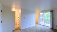 This single-floor residence features an open living area finished with neutral wall-to-wall carpeting and crisp white walls, creating a clean and versatile backdrop. The living space is well-lit by natural light streaming through a sliding glass door, which provides direct access to an outdoor area and frames a view of surrounding greenery and brick exterior detailing. The sliding door is fitted with a vertical blind track, allowing for adjustable light control and privacy.

A wide opening connects the living area to an interior hallway, which leads to the kitchen and additional spaces beyond. The kitchen, visible from the living area, features light cabinetry and appears to include standard appliances, illuminated by warm overhead lighting. The hallway further extends to what appears to be additional rooms within the unit.

Smooth, light-toned ceilings run throughout, contributing to an airy and open feel. Electrical outlets and a thermostat are positioned along the walls, reflecting practical, functional design. The overall layout is straightforward and efficient, making the space adaptable for a variety of furnishing arrangements.