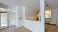 This single-floor space is designed with a clean, open layout that emphasizes light and flow. The main living area is anchored by a set of elegant French doors fitted with gridded glass panels and transom windows above, allowing natural light to pour into the interior and creating an inviting entryway. Vaulted ceilings rise above the space, adding a sense of openness and architectural interest, while a globe pendant light fixture hangs as a simple, functional accent. The walls are finished in crisp white throughout, contributing to the bright, airy atmosphere. Neutral-toned wall-to-wall carpeting runs underfoot, providing a soft and cohesive base across the living areas. A defining architectural feature is the half-wall pass-through that connects the main living space to the kitchen beyond, offering a glimpse of warm wood cabinetry and pendant lighting in the adjacent room. This open sightline promotes a connected feel between spaces. Built-in HVAC vents are integrated neatly into the walls, and a wall-mounted thermostat provides convenient climate control. Crown molding details add a subtle finishing touch to the overall design. The space presents a move-in ready, functional layout with classic architectural details throughout.