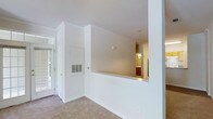 This single-floor space is designed with a clean, open layout that emphasizes light and flow. The main living area is anchored by a set of elegant French doors fitted with gridded glass panels and transom windows above, allowing natural light to pour into the interior and creating an inviting entryway. Vaulted ceilings rise above the space, adding a sense of openness and architectural interest, while a globe pendant light fixture hangs as a simple, functional accent. The walls are finished in crisp white throughout, contributing to the bright, airy atmosphere. Neutral-toned wall-to-wall carpeting runs underfoot, providing a soft and cohesive base across the living areas. A defining architectural feature is the half-wall pass-through that connects the main living space to the kitchen beyond, offering a glimpse of warm wood cabinetry and pendant lighting in the adjacent room. This open sightline promotes a connected feel between spaces. Built-in HVAC vents are integrated neatly into the walls, and a wall-mounted thermostat provides convenient climate control. Crown molding details add a subtle finishing touch to the overall design. The space presents a move-in ready, functional layout with classic architectural details throughout.