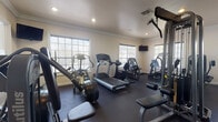 This fitness center offers a thoughtfully organized workout space designed to support a full range of exercise routines. The room features dark rubber flooring throughout, providing durability and traction across the entire workout area. Crown molding and neutral-toned walls give the space a polished, finished aesthetic that sets it apart from a typical gym environment.

Natural light flows in generously through multiple large, grid-paned windows positioned along the walls, creating a bright and inviting atmosphere. Recessed ceiling lighting supplements the natural light, ensuring the space remains well-lit at all hours. A wall-mounted flat-screen television is positioned for easy viewing during cardio sessions.

The cardio section is well-appointed with treadmills, an elliptical trainer, and a rowing machine, offering users a variety of aerobic workout options. The strength-training area includes multiple cable and resistance machines targeting different muscle groups, including leg press, lat pulldown, and cable pulley systems, among others. A large floor-to-ceiling mirror spans one wall, enhancing the sense of openness and allowing users to monitor their form during exercise.

The space is accessible via a glass-paned door and includes a clearly marked emergency exit, reflecting attention to safety. Overall, this fitness center combines functional equipment with a clean, well-maintained environment suitable for a comprehensive daily workout routine.