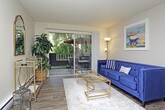 This single-floor residence is designed with a cohesive, coastal-inspired aesthetic that flows seamlessly from room to room. The living area serves as the heart of the space, anchored by rich hardwood-style flooring and neutral walls that provide a warm backdrop for vibrant decor. A bold navy velvet sofa with patterned throw pillows takes center stage, complemented by metallic gold accent furniture, an open decorative bookshelf, and a sliding glass door that invites natural light into the room. Coastal artwork and sculptural wall decor add personality and visual interest throughout.

The kitchen is efficiently laid out with white cabinetry, granite-look countertops, and white appliances, including a coil-burner range and a full-size refrigerator. Charming floral wall accents add a playful touch to the cooking space.

The bedroom offers a serene retreat, featuring a tufted upholstered headboard, mirrored nightstands, and coordinating table lamps that create a soft, restful ambiance. A multi-panel monochromatic seascape canvas above the bed adds a sophisticated focal point.

The bathroom is clean and practical, with a full tub-and-shower combination, wood-style flooring, and simple, tasteful wall accents. A textured ceiling is present throughout the home, tying together the overall design character of this well-appointed space.