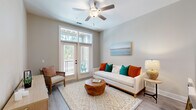 Located in Charleston, SC, this thoughtfully designed one-bedroom, one-bathroom residence offers a well-appointed single-floor layout with a cohesive, modern aesthetic throughout. Light wood-look flooring flows through the main living areas, complemented by neutral warm-gray walls and crisp white trim that create a bright, welcoming ambiance.

The open-concept kitchen features contemporary flat-front cabinetry in a light wood tone, stone-look quartz countertops, stainless steel appliances including a range, microwave, dishwasher, and refrigerator, and a breakfast bar with pendant lighting — ideal for casual dining. The kitchen opens directly to the living area, which is anchored by a ceiling fan and french doors that provide both natural light and access to a private balcony with outdoor views.

The bedroom offers plush carpeting, generous natural light from dual windows with sheer curtains, and a comfortable, calming atmosphere. A dedicated closet provides practical storage. The full bathroom includes a wide vanity with stone-look countertops, modern hardware, and wood-toned cabinetry. A dedicated laundry closet houses a stacked washer and dryer unit, adding everyday convenience. Two hallways connect the space efficiently, making the layout feel organized and functional throughout.