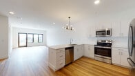 This contemporary single-floor residence showcases a clean, modern design with an open-concept layout that seamlessly connects the kitchen and living area. Light wood flooring runs throughout the space, lending warmth and continuity to every room.

The kitchen is a standout feature, equipped with white shaker-style cabinetry, marble-look quartz countertops, a classic subway tile backsplash, and a full suite of stainless steel appliances including a French door refrigerator, electric range, over-the-range microwave, and dishwasher. A peninsula with an integrated sink and built-in storage creates a natural division between the kitchen and the expansive living area, which is accented by a stylish multi-arm chandelier and large windows that invite abundant natural light.

The bedroom is bright and airy, featuring a walk-in closet and a sun-filled window with outdoor views. The bathroom is finished with large-format light gray tile surrounding a soaking tub and shower combination, paired with chrome fixtures and wood-look flooring. In-unit laundry is also included. Throughout, crisp white walls and recessed lighting contribute to the space's fresh, contemporary ambiance.