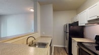 This 1-bedroom, 1-bathroom unit is designed for comfortable, functional living across a single floor. The open living area features soft carpeting, a ceiling fan, and a sliding glass door that leads to a private balcony with outdoor views, filling the space with natural light. The entryway transitions seamlessly from tile to carpet, with multiple doors providing access to closets and adjoining rooms.

The kitchen is efficiently laid out with a pass-through counter connecting it to the living area. It features white cabinetry, granite-style countertops, a stainless steel sink, wood-look flooring, and stainless steel appliances including a refrigerator and electric range, complemented by an overhead ventilation hood.

The bedroom is a comfortable, carpeted retreat with a large window overlooking lush greenery, offering privacy and natural light. Neutral wall tones and a textured ceiling carry throughout the unit, creating a cohesive, understated aesthetic.

The bathroom is clean and well-appointed, featuring a single-sink vanity with matching granite-style countertops, white cabinetry, a full tub-shower combination with white tile accents and a terracotta tile border, and wood-look flooring that ties seamlessly to the rest of the unit.