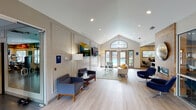 This single-story clubhouse is designed with a contemporary aesthetic, offering residents a welcoming and functional communal environment. The main lounge is the centerpiece of the space, characterized by soaring vaulted ceilings with recessed lighting and light hardwood-style flooring that runs throughout. An arched transom window crowns a set of French doors that open directly toward an outdoor pool area, flooding the interior with natural light and providing a seamless indoor-outdoor connection. The lounge is furnished with a mix of modern navy swivel chairs, a neutral-toned sofa with accent pillows, and glass coffee tables, all arranged to encourage comfortable gathering. A wood-paneled accent wall with a mounted circular metallic art piece anchors one side of the room, while large flat-screen televisions are mounted along the opposite wall for entertainment. Adjacent to the lounge is a well-appointed kitchenette, distinguished by its bold penny-tile backsplash, white cabinetry, quartz countertops, stainless steel appliances including a refrigerator, dishwasher, and microwave, open wood shelving, and a beverage cooler. A fitness center is also part of the facility, equipped with a range of exercise machines and free weights to support an active lifestyle.