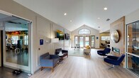 This single-story clubhouse is designed with a contemporary aesthetic, offering residents a welcoming and functional communal environment. The main lounge is the centerpiece of the space, characterized by soaring vaulted ceilings with recessed lighting and light hardwood-style flooring that runs throughout. An arched transom window crowns a set of French doors that open directly toward an outdoor pool area, flooding the interior with natural light and providing a seamless indoor-outdoor connection. The lounge is furnished with a mix of modern navy swivel chairs, a neutral-toned sofa with accent pillows, and glass coffee tables, all arranged to encourage comfortable gathering. A wood-paneled accent wall with a mounted circular metallic art piece anchors one side of the room, while large flat-screen televisions are mounted along the opposite wall for entertainment. Adjacent to the lounge is a well-appointed kitchenette, distinguished by its bold penny-tile backsplash, white cabinetry, quartz countertops, stainless steel appliances including a refrigerator, dishwasher, and microwave, open wood shelving, and a beverage cooler. A fitness center is also part of the facility, equipped with a range of exercise machines and free weights to support an active lifestyle.