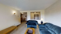 This single-floor residence offers a practical and comfortable layout designed for everyday living. The living room serves as the central gathering space, featuring wall-to-wall carpeting, a textured ceiling, and soft, neutral-toned walls that create a warm and inviting atmosphere. The room provides ample space for seating arrangements and is illuminated by wall-mounted light fixtures that add a cozy ambiance.

A pass-through opening in the wall connects the living area to an adjacent space — visible through the opening is a closet or entryway area, complete with a door and overhead lighting, offering convenient storage access. To the left, an open doorway leads directly into the dining area, which features hardwood-style flooring and a dining table with chairs, providing a distinct yet seamlessly connected space for meals.

The overall layout of the home promotes an easy flow between the living, dining, and entry areas, making the space feel open and well-connected. The consistent use of neutral wall colors and understated finishes throughout creates a cohesive and clean aesthetic. The single-floor design ensures accessibility and ease of movement throughout the entire home.