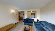 This single-floor residence offers a practical and comfortable layout designed for everyday living. The living room serves as the central gathering space, featuring wall-to-wall carpeting, a textured ceiling, and soft, neutral-toned walls that create a warm and inviting atmosphere. The room provides ample space for seating arrangements and is illuminated by wall-mounted light fixtures that add a cozy ambiance.

A pass-through opening in the wall connects the living area to an adjacent space — visible through the opening is a closet or entryway area, complete with a door and overhead lighting, offering convenient storage access. To the left, an open doorway leads directly into the dining area, which features hardwood-style flooring and a dining table with chairs, providing a distinct yet seamlessly connected space for meals.

The overall layout of the home promotes an easy flow between the living, dining, and entry areas, making the space feel open and well-connected. The consistent use of neutral wall colors and understated finishes throughout creates a cohesive and clean aesthetic. The single-floor design ensures accessibility and ease of movement throughout the entire home.