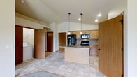 This single-floor space showcases a thoughtfully designed open-concept layout that seamlessly blends the kitchen and living areas into a cohesive, functional environment. The kitchen is a standout feature, highlighted by warm honey-toned wood cabinetry that provides ample storage, granite countertops with subtle veining, and a peninsula island that offers additional prep space and casual seating. Black appliances, including a refrigerator, range, and built-in microwave, add a sleek, modern contrast to the lighter cabinetry and countertops. Two pendant lights hang gracefully above the island, providing focused task lighting and an elegant design touch. Recessed lighting throughout the kitchen ensures the space is well-illuminated. The vaulted ceilings rise dramatically overhead, creating an expansive, airy atmosphere that enhances the sense of openness. Light tile flooring extends throughout the main living area, lending a clean and cohesive look underfoot. Adjacent to the kitchen, a hallway leads to additional rooms, with warm wood doors maintaining a consistent design palette. Overall, the space offers a welcoming and practical environment with a neutral, timeless aesthetic.