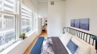 Located at 173–191 S Wabash Ave in Chicago's iconic Loop neighborhood, this single-floor residence blends industrial character with contemporary design. Soaring ceilings throughout create a dramatic sense of volume, while herringbone-patterned hardwood floors add warmth and visual interest to the open layout.

The living area is anchored by a bold slate-blue accent wall and features expansive vertical space, making the room feel airy and inviting. A sectional sofa, organic wood coffee table, and curated wall art contribute to a modern yet comfortable aesthetic.

The kitchen is efficiently designed with light wood cabinetry, white quartz countertops, open shelving, and a full suite of stainless steel appliances including a gas range, built-in microwave, dishwasher, and refrigerator. A gold-toned faucet adds a refined touch.

The bedroom benefits from a bank of large windows that flood the space with natural light and offer views of the surrounding Chicago cityscape. High ceilings continue here, reinforcing the loft-like character of the unit.

The bathroom is clean and modern, featuring a white vanity with brass hardware, a large mirror, and a neutral gray floor, providing a functional and polished finish. The overall ambiance of this space reflects a thoughtfully curated urban loft aesthetic in the heart of downtown Chicago.