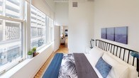 Located at 173–191 S Wabash Ave in Chicago's iconic Loop neighborhood, this single-floor residence blends industrial character with contemporary design. Soaring ceilings throughout create a dramatic sense of volume, while herringbone-patterned hardwood floors add warmth and visual interest to the open layout.

The living area is anchored by a bold slate-blue accent wall and features expansive vertical space, making the room feel airy and inviting. A sectional sofa, organic wood coffee table, and curated wall art contribute to a modern yet comfortable aesthetic.

The kitchen is efficiently designed with light wood cabinetry, white quartz countertops, open shelving, and a full suite of stainless steel appliances including a gas range, built-in microwave, dishwasher, and refrigerator. A gold-toned faucet adds a refined touch.

The bedroom benefits from a bank of large windows that flood the space with natural light and offer views of the surrounding Chicago cityscape. High ceilings continue here, reinforcing the loft-like character of the unit.

The bathroom is clean and modern, featuring a white vanity with brass hardware, a large mirror, and a neutral gray floor, providing a functional and polished finish. The overall ambiance of this space reflects a thoughtfully curated urban loft aesthetic in the heart of downtown Chicago.