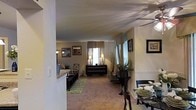 This single-story residence showcases an open-concept layout that seamlessly connects the living and dining areas, creating a cohesive and functional flow throughout the main living space. The living area is finished with plush, neutral-toned carpeting that adds warmth underfoot, while textured ceilings contribute character to the interior. Multiple windows dressed with soft fabric valances allow natural light to filter into the space, brightening the warm, neutral color palette found throughout. A dark-finished console table positioned beneath the windows serves as a natural focal point in the living area, complemented by a pair of coordinating table lamps that provide ambient lighting. Wall-mounted mirrors and framed artwork enhance the sense of depth and visual interest along the walls. A decorative area rug anchors the living space, while lush potted plants introduce organic texture and life to the room. The dining area, partially visible from the main living space, features a dark wood dining table and chairs set with place settings, suggesting a welcoming environment for entertaining. A half-wall with a light-toned countertop separates and subtly defines the dining zone, adding an architectural detail that contributes to the overall layout. Framed wall art completes the polished, cohesive aesthetic.