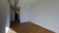 This single-floor space blends industrial character with clean, modern finishes to create an open and functional environment. The main living area is defined by rich, wide-plank hardwood flooring that runs throughout, adding warmth and a sense of continuity to the space. Crisp white walls provide a neutral backdrop, while the ceiling features an exposed metal ventilation duct that lends an industrial loft-like aesthetic to the interior.

Ceiling-mounted lighting fixtures are strategically placed to illuminate the space, including a central flush-mount light in the main area and a pendant cluster visible along the hallway corridor, adding layers of light and visual interest. The hallway, partially visible from the main room, leads further into the unit and is lined with white doors, hinting at additional rooms beyond.

The layout feels open and unencumbered, allowing for flexible furniture arrangements and versatile use of the space. The combination of industrial details—such as the exposed ductwork and utilitarian fixtures—alongside polished hardwood floors and smooth white walls creates a balanced, contemporary atmosphere suited to a variety of uses.