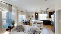 This 2-bedroom, 2-bathroom single-floor residence offers a thoughtfully designed open-concept layout that seamlessly connects the living, dining, and kitchen areas. The living room is anchored by a plush, light-toned sofa accented with bold black-and-white patterned throw pillows, creating a clean and contemporary aesthetic. Natural light pours in through multiple windows dressed with sheer white curtains and complementary window treatments, enhancing the bright and airy atmosphere throughout.

The kitchen is a standout feature, showcasing dark cabinetry that contrasts elegantly with a white countertop island. Pendant lighting in brushed metal finishes hangs above the island, which provides bar-style seating with sleek industrial stools — ideal for casual dining or entertaining. Track lighting runs throughout the main living areas, ensuring versatile and well-distributed illumination.

The dining area sits adjacent to the kitchen and living room, furnished with a dark rectangular table and coordinating chairs beneath a cluster of decorative wall art. Light wood-toned flooring flows continuously across the open floor plan, unifying the spaces with warmth and cohesion. The overall ambiance is modern, polished, and welcoming.
