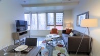 This stylish single-floor urban residence offers a thoughtfully designed open-concept layout that seamlessly blends living and dining spaces. The living area is anchored by a large, multi-panel window that floods the room with natural light and frames impressive city skyline views, creating an energizing connection to the surrounding urban environment. A ceiling fan adds a practical touch overhead.

The living space is comfortably appointed with a plush grey sofa accented by warm rust-toned throw pillows, a modern accent chair in a rich terracotta hue, and a round glass-topped coffee table resting on a vibrant, multi-colored area rug. A sleek media console with a wall-mounted flat-screen television anchors one side of the room, while a tasteful framed artwork adds character to the walls.

Adjacent to the living area, the dining space features a contemporary wooden table set with place settings, creating an inviting area for meals. The neutral color palette throughout — soft whites, warm greys, and earthy accent tones — contributes to a cohesive and welcoming ambiance. The combination of modern furnishings, abundant natural light, and panoramic city views makes this space a compelling urban retreat.